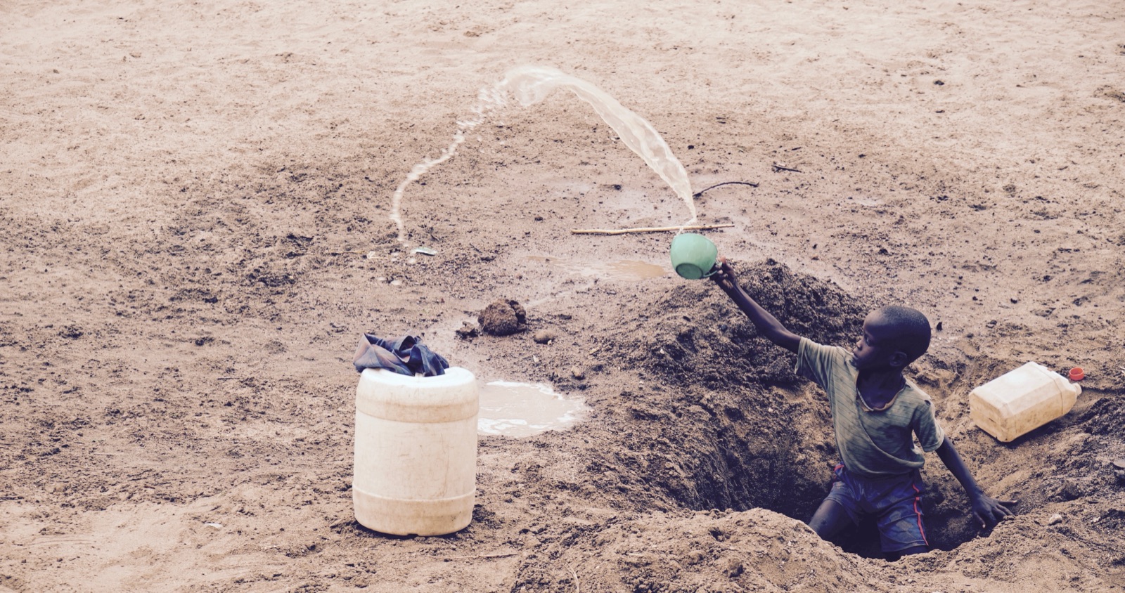 A boy in a shallow well pours water into a plastic drum.